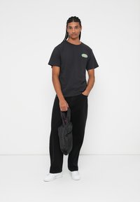 Sort bomulds T-shirt med rund halsudskæring og lille farverig grafik på brystet, parret med løstsiddende sorte bukser og hvide sneakers.
