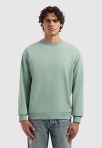 Jonge man met krullend haar, gekleed in een lichtgroene sweatshirt en vervaagde blauwe jeans, staande met een neutrale uitstraling tegen een effen achtergrond.