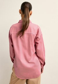 Camisa de gran tamaño en color rosa, hecha de algodón, con mangas largas, dobladillo curvado y detalles de botones en los puños. Diseño liso, sin patrones.