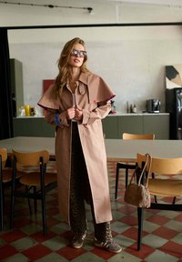 Kobieta w okularach przeciwsłonecznych, ubrana w beżowy trencz i spodnie w leopardzi wzór, stoi w pokoju z stołem, krzesłami i torebką na jednym z krzeseł.