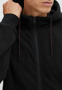 Schwarzer Fleece-Hoodie mit durchgehendem Reißverschluss, verstellbarem Kapuzen und kontrastierenden roten Kordelzügen. Weicher Stoff und taillierte Passform.
