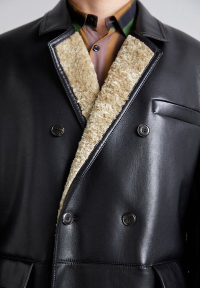 Black leather double-breasted coat with a plush beige fur collar, featuring two button closures and a chest pocket. Smooth texture.