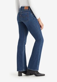 Femme portant un jean bootcut bleu foncé, un haut blanc à manches longues et des bottines noires à talons, debout de profil.