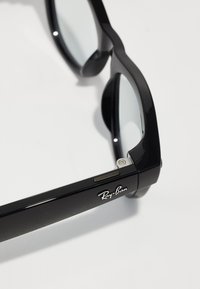 Schwarze Kunststoff-Sonnenbrille mit klarem Glas, versehen mit einer glatten Oberfläche, abgerundeten Kanten und einem Metallelement nahe dem Scharnier mit einem Logo.