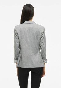 Donna con lunghi capelli scuri, che indossa un blazer grigio con i risvolti arrotolati e pantaloni neri, in piedi di spalle contro uno sfondo bianco.