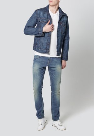 Jeansjacka i mörkblått med knappstängning och två bröstfickor, bärs över en vit t-shirt. Slim-fit ljusblå jeans och vita sneakers.