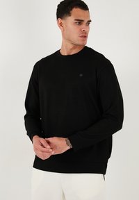 Sudadera negra de manga larga, diseño de cuello redondo y un pequeño detalle de logo. La tela parece suave, con un corte holgado y puños acanalados.