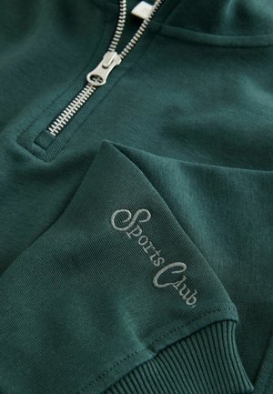Tamsiai žalias užtraukiamas džemperis, pagamintas iš minkštos medžiagos. Rodo sidabrinį užtrauktuką ir siuvinėtą "Sporto Klubas" užrašą ant rankovės.