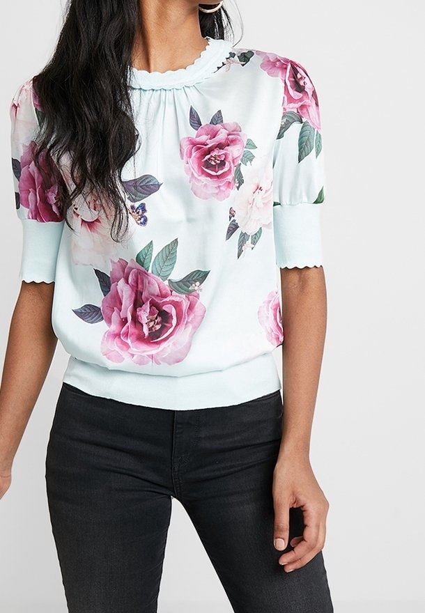 Femme portant un chemisier bleu clair à imprimés de roses roses et blanches, avec des manches bouffantes, associé à un jean noir.