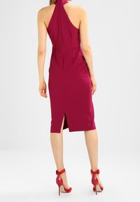 Figurbetontes magentafarbenes Kleid mit hohem Kragen und offenem Rücken, ausgestattet mit einem seitlichen Schlitz. Kombiniert mit roten High Heels und Knöchelriemchen.
