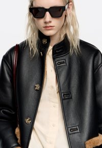 Persona que lleva una chaqueta de cuero negro oversized con grandes botones, camisa beige, gafas de sol negras y un bolso de cuero rojo oscuro.