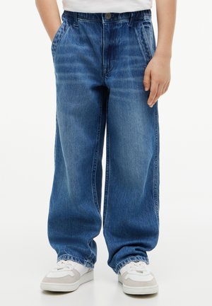 Kind trägt lockere blaue Jeans und weiße Sneakers, steht mit einer Hand teilweise in der Tasche vor einfachem Hintergrund.