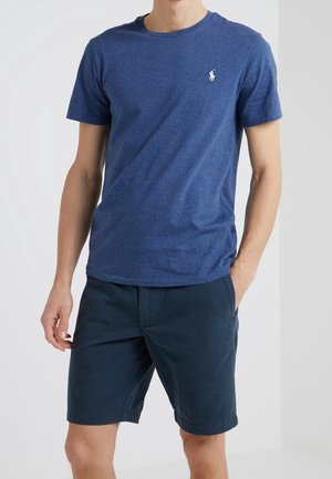 Homme portant un t-shirt bleu à manches courtes et col rond avec un petit logo blanc, ainsi qu'un short bleu marine atteignant les genoux, une main dans la poche.