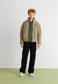 Beige fleece jacket with a zip front and two pockets, layered over a dark green hoodie, paired with black pants and grey sneakers.