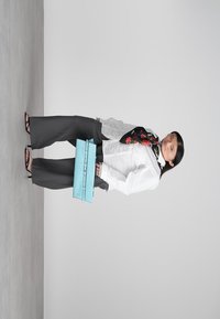 Γυναίκα με μαύρο παντελόνι φάρδος, λευκό πουκάμισο, ανθισμένο μαντήλι, μαύρες γόβες, κρατώντας μια μεγάλη μπλε τσάντα φάκελο, που στέκεται μπροστά από απλό φόντο.