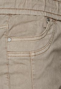 Denim beige, primer plano de los detalles del bolsillo. Acentos cosidos, botón de metal plateado y tejido texturizado. Tejidos verticales y diagonales visibles.