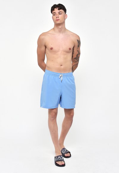 Pantaloni da bagno azzurri chiari con vita regolabile; abbinati a ciabatte scure con logo bianco. Il modello è in piedi contro uno sfondo neutro.