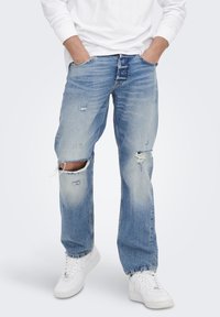 Jeans in denim azzurro chiaro con dettagli strappati, caratterizzati da ginocchia strappate, gamba dritta e una vestibilità rilassata. Indossati con sneakers bianche.