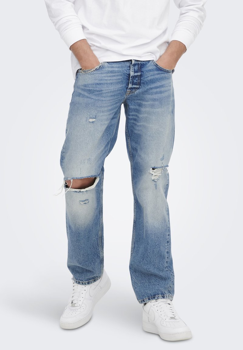 Lichtblauwe denim jeans met verwerkte details, voorzien van gescheurde knieën, rechte pijpen en een relaxte pasvorm. Draagt met witte sneakers.