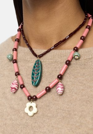 Dwa koralikowe naszyjniki na beżowym swetrze; jeden ma zielony zawieszkę w kształcie liścia, drugi różowe koraliki z zawieszkami w kształcie kwiatów i muszli.