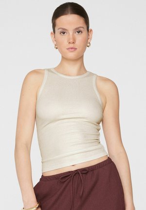 Giovane donna con capelli castani indossa un top beige smanicato a costine e pantaloni bordeaux con coulisse, posa davanti a uno sfondo chiaro e uniforme.