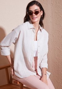Light textured button-up shirt in white with a relaxed fit. Paired with a white tank top and pink fitted pants, seated on a wooden chair.