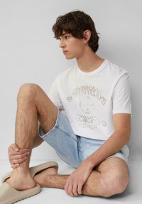 Weißes T-Shirt mit kurzen Ärmeln und hellem grafischen Text, kombiniert mit hellblauen Denim-Shorts. Model trägt beige Slip-On-Schuhe und befindet sich in sitzender Position.