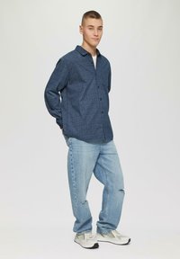 Chemise à boutons bleue à motif associée à un jean en denim clair et des chaussures de sport grises. La chemise a un col et des manches longues.