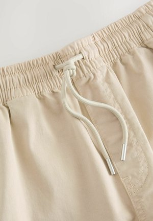 Beige elastic-waist shorts with a drawstring. Fabric is smooth and lightweight, featuring silver aglets on the cord.