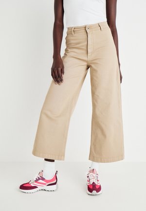 Modelo vistiendo pantalones anchos recortados beige, calcetines blancos y zapatillas rosas, rojas y blancas sobre un fondo liso.