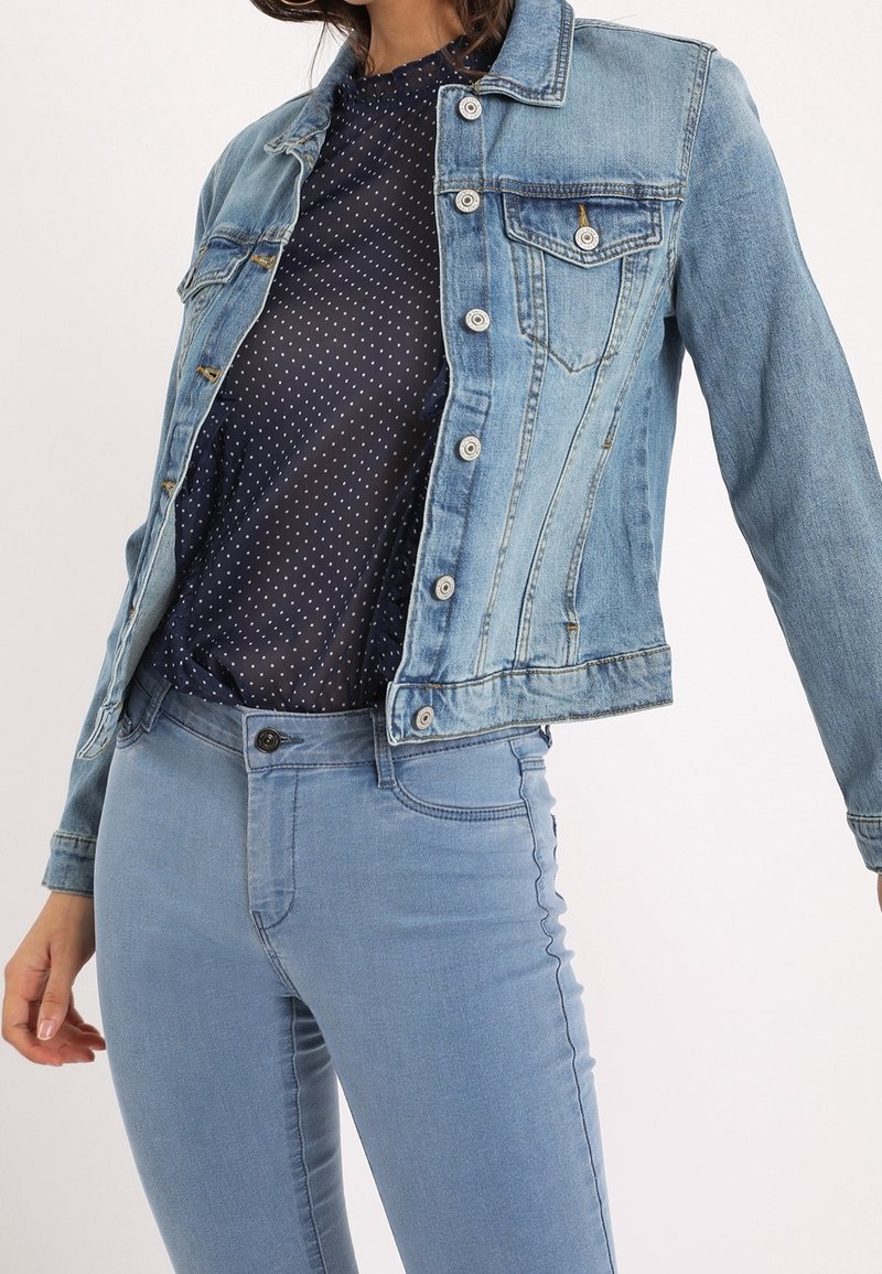 Giacca di jeans azzurro chiaro con tasche frontali, bottoni argentati e finitura leggermente slavata, abbinata a una camicia scura a pois e jeans azzurri.