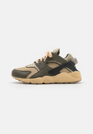 Sportieve sneaker met beige geweven stof, olijfgroene overlays, perzikkleurige veters, dikke beige zool en zwart hielbanddetail, op witte achtergrond.