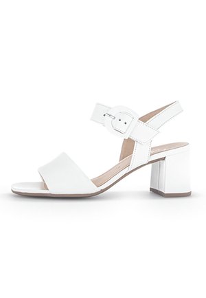 Hvide åbne sandaler med blokhæle og justerbar ankelrem, der har en rund spænde, på en hvid baggrund.