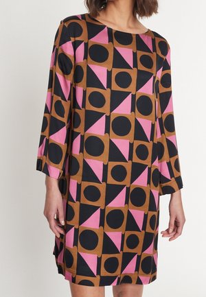 Kobieta ubrana w sukienkę do kolan z długimi rękawami, ozdobioną geometrycznym wzorem czarnych kół, brązowych kwadratów i różowych trójkątów.