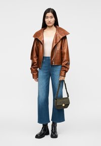 Kvinde stående iført en brun oversized læderjakke, beige top, blå vidde-benet jeans, sorte støvler og holder en olivengrøn håndtaske.