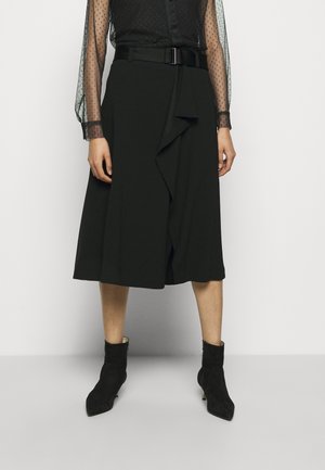 Kobieta ubrana w czarną spódnicę do kolan z falbaną z przodu, szeroki pasek, przejrzystą czarną bluzkę z długim rękawem oraz czarne botki za kostkę.