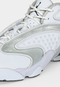 Λευκό και ασημί αθλητικό παπούτσι με μαύρο λογότυπο στη γλώσσα, λευκά κορδόνια και διάτρητο πλαϊνό πάνελ σε λευκή σόλα.