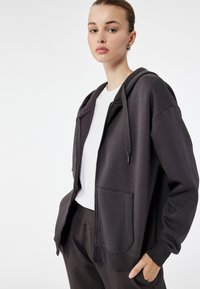 Donkergrijze zip-up hoodie gemaakt van zachte stof, met een capuchon, voorzakken en geribde manchetten. Draagt boven een wit t-shirt.