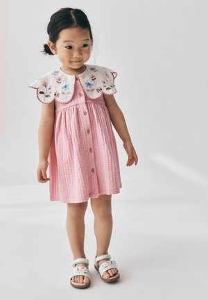 Young girl with dark hair in low buns wears a pink dress with large white floral collar and white floral sandals, standing on a plain background.