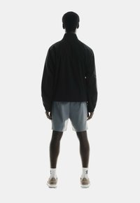 Black lightweight jacket, short gray shorts, and beige sneakers. Model's back facing camera against a white background.