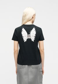 Γυναίκα με μακριά ξανθιά μαλλιά που φοράει μαύρο T-shirt με λευκά γραφικά φτερά αγγέλου και αθλητικά παπούτσια στην πλάτη, σε συνδυασμό με γκρι φούστα.