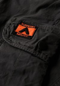 Czarna tkaninowa kieszeń z naszywką z pomarańczowym haftowanym logo z napisem "SUPERDRY" i wzorem trójkąta, zapięta okrągłym czarnym guzikiem.
