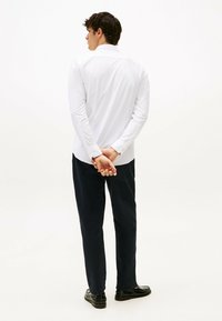 Camicia bianca a maniche lunghe con polsini a bottoni, infilata in pantaloni blu scuro. Il modello sta in piedi con le mani incrociate dietro la schiena contro uno sfondo minimale.