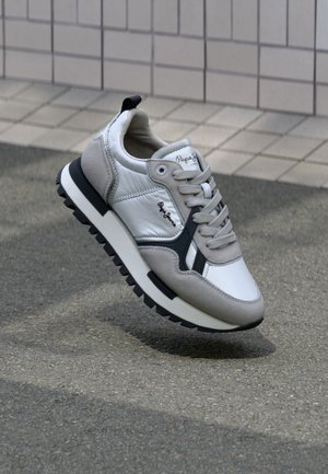 Zapatilla blanca y gris con materiales sintéticos y de ante, puntera redonda, acentos negros contrastantes, suela texturizada y detalle del logo en el costado.