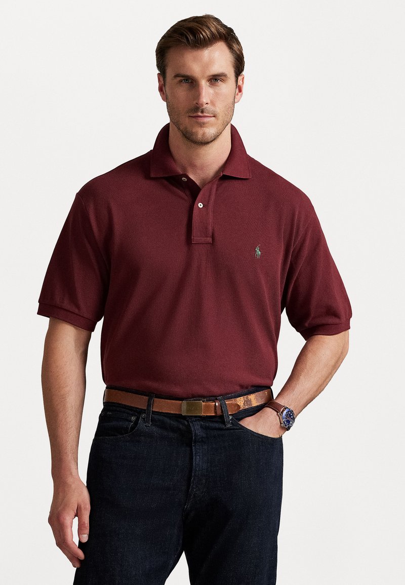 Burgundowa koszulka polo z klasycznym kołnierzykiem, dwoma guzikami i zielonym logo po lewej stronie klatki piersiowej, w połączeniu z ciemnymi dżinsami i brązowym paskiem.