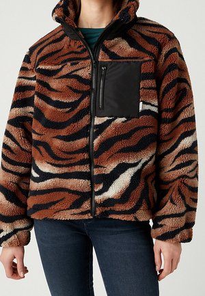 Personne portant une veste polaire à rayures tigre marron et noire, avec une fermeture éclair frontale et une poche poitrine zippée noire.