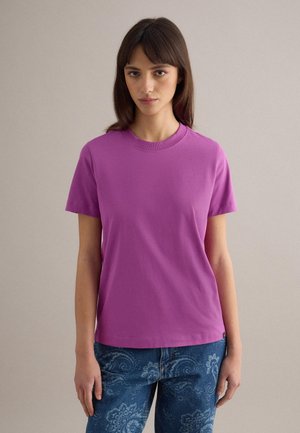 Giovane donna con capelli castani lisci, che indossa una t-shirt viola a maniche corte e jeans blu con motivo paisley, in piedi davanti a uno sfondo semplice.