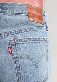 Jeans de mezclilla azul claro con un parche de cuero de marca y una etiqueta roja. Presenta bolsillos traseros con detalles de costura y un ajuste clásico.