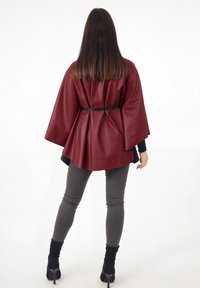 Capa extragrande de cuero sintético burdeos con un cinturón negro, que presenta un acabado brillante, mangas anchas y una silueta relajada. Jeans ajustados grises completan el look.