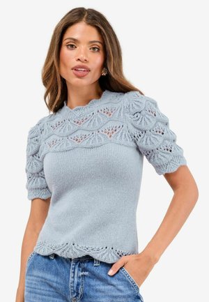 Frau, die einen hellblauen, gestrickten Kurzarmpullover mit Spitzenmustern trägt, kombiniert mit blauen Jeans, und mit einer Hand in der Tasche posiert.